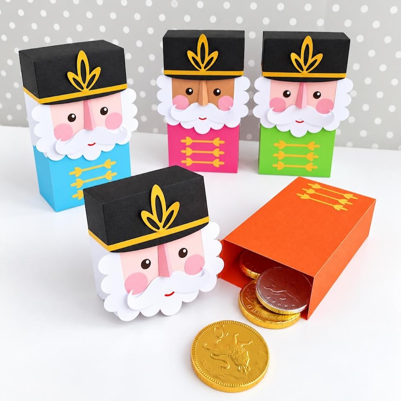 Op de afbeelding: Vier papieren notenkraker-cadeaudoosjes in blauw, roze, groen en oranje, elk met een zwarte hoed met geel embleem, witte baard en roze wangen. Een oranje doosje ligt open en toont gouden en zilveren chocolademunten aan de binnenkant.