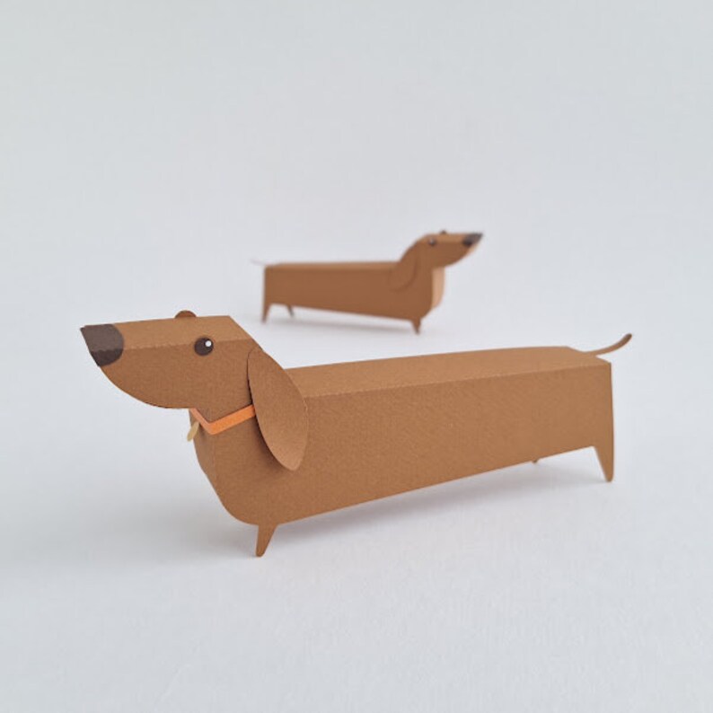May include: Two brown paper dachshund figurines. The dachshund on the left is facing the viewer and has a small orange collar. The dachshund on the right is facing away from the viewer.