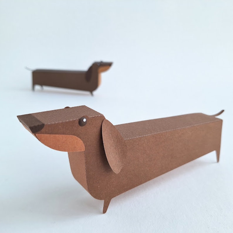 May include: Two brown paper dachshund figurines. The dachshund on the left is facing the right and the dachshund on the right is facing the left.
