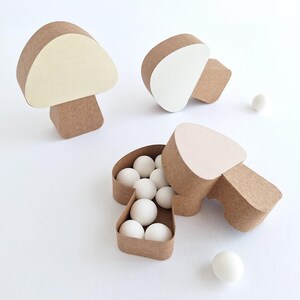 Pode incluir: Tr&ecirc;s caixas de papel castanho em forma de cogumelo. Uma caixa est&aacute; aberta e cheia de bolas de doces brancas. As outras duas caixas est&atilde;o fechadas. As caixas est&atilde;o sobre uma superf&iacute;cie branca.