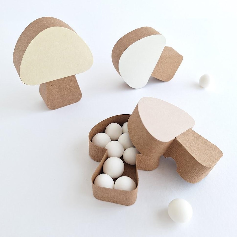 Pode incluir: Tr&ecirc;s caixas de papel castanho em forma de cogumelo. Uma caixa est&aacute; aberta e cheia de bolas de doces brancas. As outras duas caixas est&atilde;o fechadas. As caixas est&atilde;o sobre uma superf&iacute;cie branca.