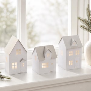 Peut inclure: Trois maisons lumineuses en papier blanc avec des toits gris clair et des fenêtres découpées. Chaque structure présente un toit à pignon, une porte d'entrée et plusieurs ouvertures carrées diffusant une lueur chaude et douce.
