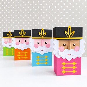 Op de afbeelding: Vier rechthoekige papieren cadeauzakjes ontworpen als notenkraker-soldaten. Elk zakje heeft een zwarte hoed met een geel embleem, een witte snor, roze wangen en een uniform in groen, oranje, blauw of roze met gele details op de voorzijde.