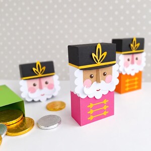 Op de afbeelding: Drie rechthoekige papieren doosjes in de vorm van notenkrakersoldaten met zwarte hoeden, witte baarden en roze of oranje uniformen. Elk doosje heeft een geel embleem op de hoed en gele horizontale lijnen op de borst. Gouden en zilveren munten liggen ernaast.