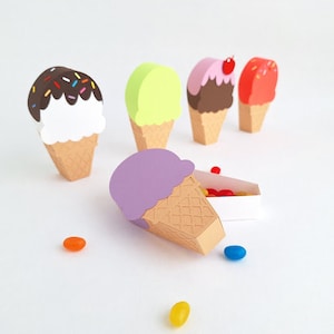 May include: A set of five ice cream cone shaped boxes, four are filled with colourful sweets and one is empty. The boxes are made of paper and are decorated with different colours and designs. The boxes are perfect for holding small gifts or treats.