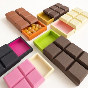 May include: A collection of rectangular sliding boxes shaped like chocolate bars, featuring various colors including brown, cream, black, pink, and green. Some boxes are open to reveal contrasting interior colors or small yellow candies.