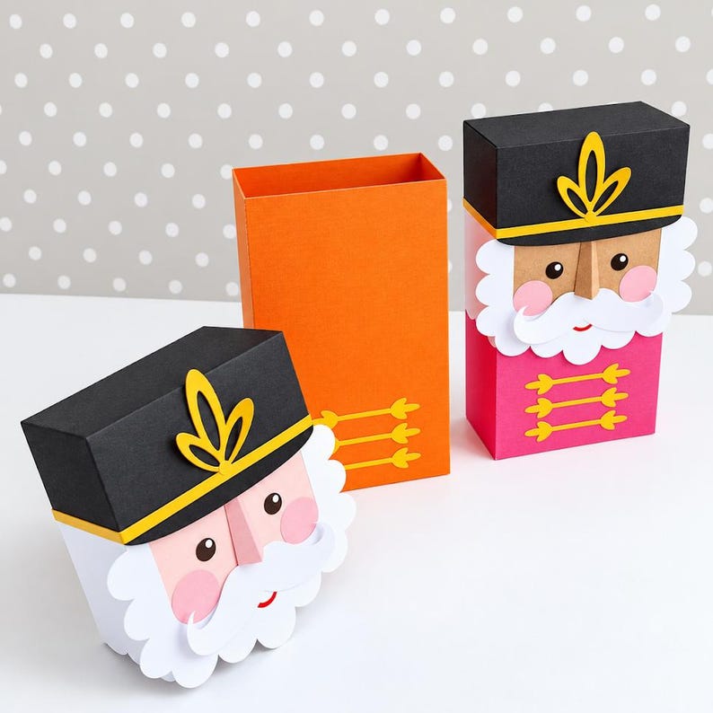 Op de afbeelding: Twee rechthoekige papieren doosjes ontworpen als notenkrakerfiguren. E&eacute;n doosje is volledig gemonteerd met een zwarte hoed, witte baard en roze jasje. Het andere doosje is gedeeltelijk gedemonteerd en toont een oranje binnenkant.