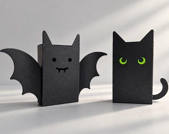 May include: Two black rectangular boxes shaped like creatures. One box features bat wings, two small circular eyes, and two fangs. The other box is shaped like a cat with a curved tail and two bright green crescent-shaped eyes.