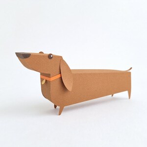 May include: A brown paper dachshund with an orange collar and a black nose. The dachshund is made of paper and has a 3D effect.