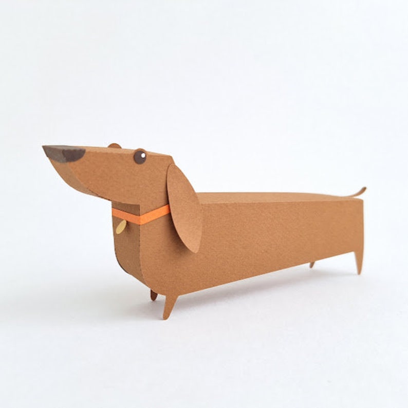 May include: A brown paper dachshund with an orange collar and a black nose. The dachshund is made of paper and has a 3D effect.