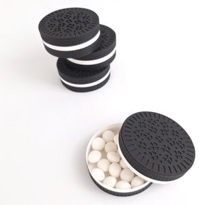 Può includere: Contenitori neri e bianchi a forma di biscotto. Un contenitore è aperto, rivelando caramelle bianche. I contenitori sono impilati e disposti su uno sfondo bianco. I coperchi hanno un motivo decorativo.