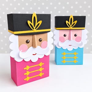 Op de afbeelding: Twee rechthoekige papieren geschenkdozen ontworpen als notenkraker-soldaten. De ene doos is roze en de andere lichtblauw. Beide hebben een zwarte hoed met een geel embleem, een witte papieren baard, roze wangen en drie horizontale gele decoratieve lijnen.