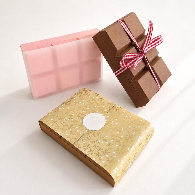 May include: Three rectangular boxes: one translucent pink box with six compartments, one brown box shaped like a chocolate bar tied with a red and white gingham ribbon, and one box wrapped in gold paper with a white floral pattern and a blank circular label.