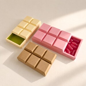 May include: Three rectangular sliding boxes shaped like chocolate bars. One box is light yellow with a green interior, one is pink containing red jelly beans, and one is brown. Each box features a raised grid pattern on the sliding lid.
