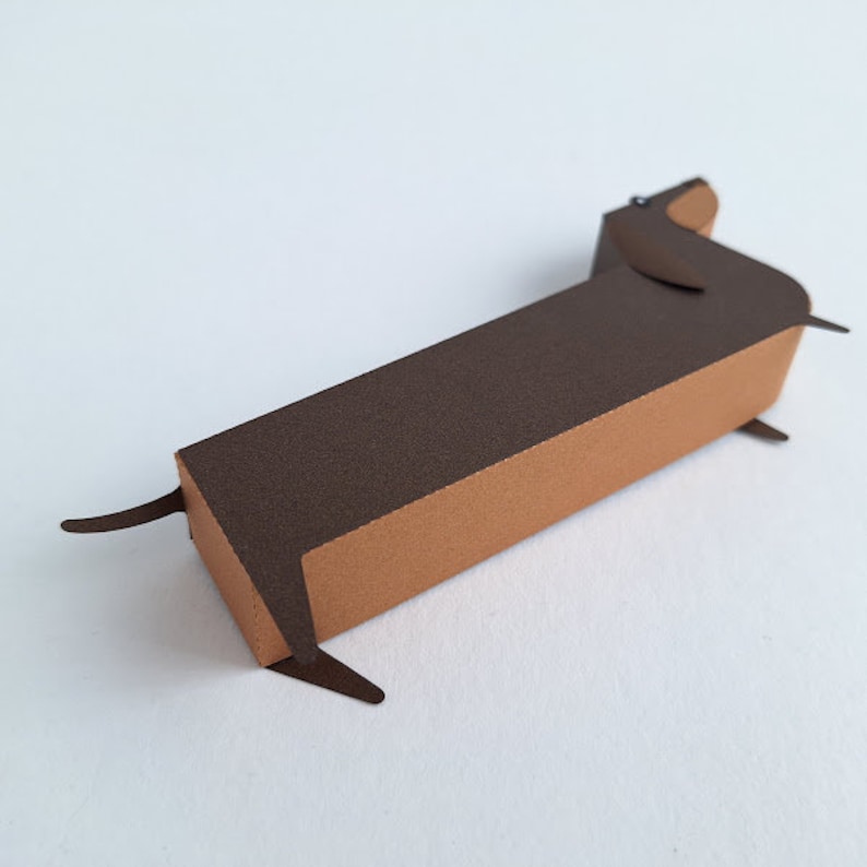 May include: A brown paper dachshund figurine with a brown and orange body. The figurine is made of paper and has a simple, minimalist design.