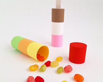 May include: A colorful paper tube with a green, orange, and yellow design, with a stick attached to the top. The tube is open and jelly beans are spilling out. There is a red cylinder and a brown, white, and pink cylinder with a stick attached to the top.