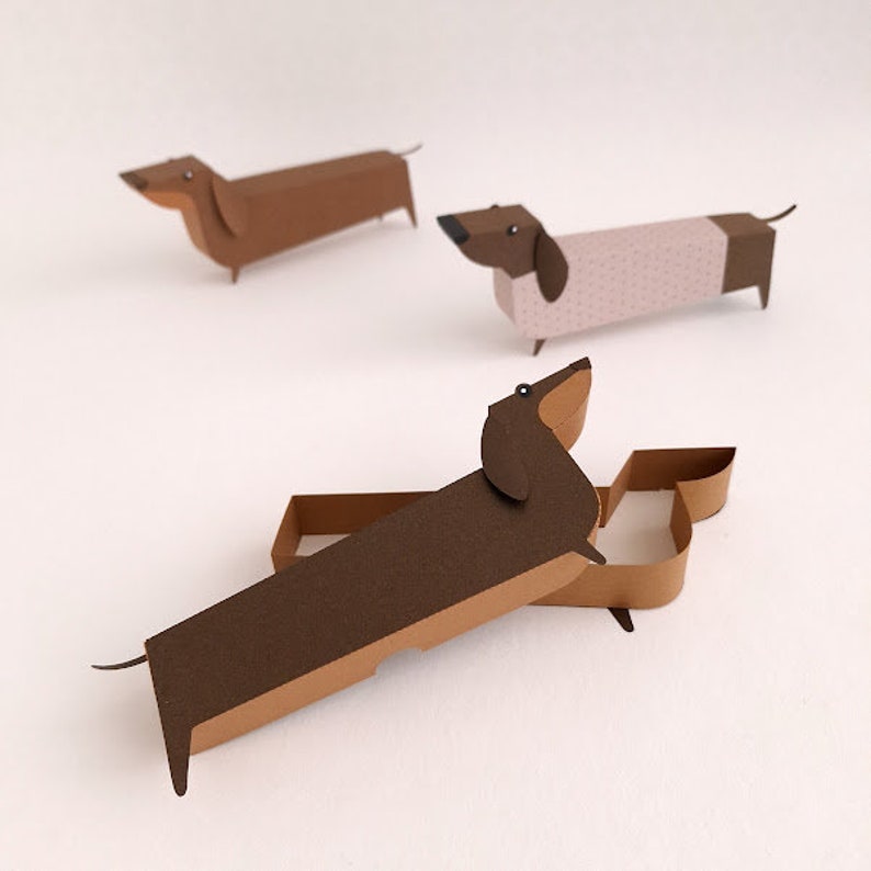 May include: Three paper dachshund dog figurines. One is brown, one is pink with white polka dots, and one is brown with a tan underside.
