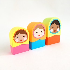 Könnte beinhalten: Drei bunte Nistpuppen mit verschiedenen Haar- und Hauttönen. Die Puppen sind aus Papier und haben einen skurrilen, cartoonartigen Stil.