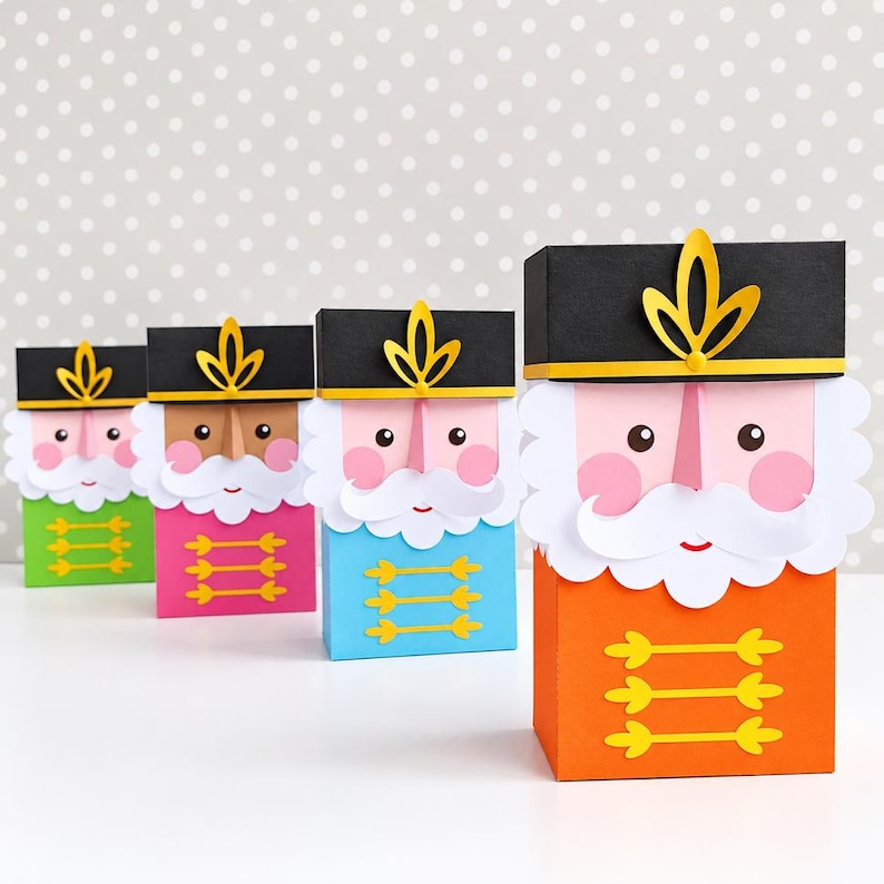 Op de afbeelding: Vier rechthoekige papieren geschenkdozen ontworpen als notenkraker-soldaten. Elke doos heeft een zwarte hoed met een geel embleem, een witte papieren baard, roze wangen en een kleurrijke basis met drie horizontale gele decoratieve lijnen.