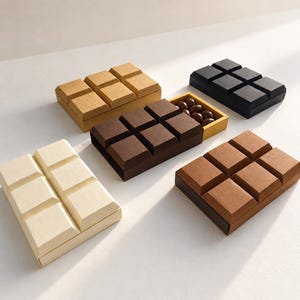 May include: Five rectangular gift boxes shaped like chocolate bars, each featuring a grid of six raised squares on the lid. The boxes are colored in cream, tan, brown, dark brown, and black. One brown box is partially open, revealing dark candies.