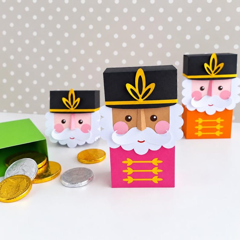 Op de afbeelding: Drie notenkraker-doosjes van papier in roze en oranje, voorzien van zwarte hoeden met gele emblemen, witte snorren en roze wangen. Een open groen doosje aan de linkerkant bevat diverse ronde chocolademunten in goud en zilver.