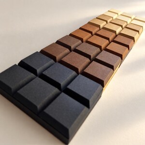 May include: A rectangular grid of square blocks arranged in three parallel rows, featuring a color gradient that transitions from dark charcoal gray at the front to progressively lighter shades of brown, tan, and cream toward the back.