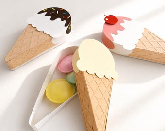 May include: Three ice cream cone-shaped paper boxes. One is closed with chocolate frosting and sprinkles, one is closed with strawberry frosting and a cherry, and one is open, revealing small, round, pastel-colored items inside.