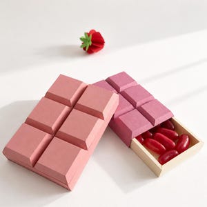 May include: Two rectangular boxes shaped like chocolate bars with raised square segments. One box is light pink, and the other is a darker magenta with a sliding wooden tray containing several shiny red jelly bean candies. A small red paper strawberry.