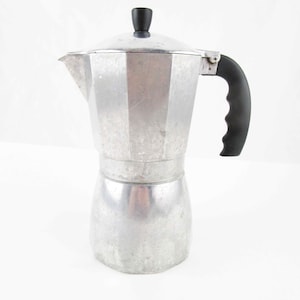 May include: A silver-colored aluminum stovetop espresso maker with an octagonal base and upper chamber. It features a black plastic handle with a contoured grip and a matching black knob on the hinged lid. The metal surface shows a matte finish.