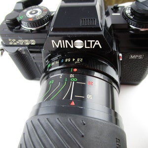 May include: A black Minolta X-700 film camera with a black lens. The camera has a silver label that reads "Minolta" and "X-700". The lens has a black rubber grip and a silver ring with markings for aperture settings.