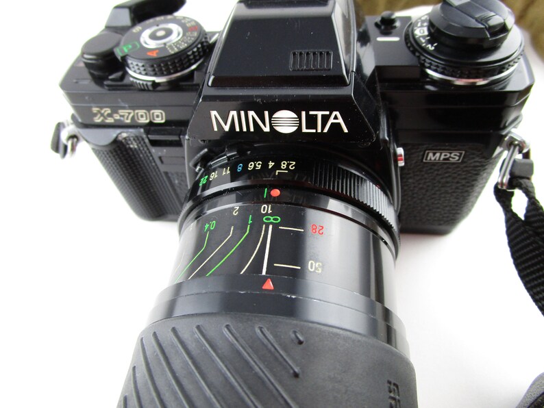 May include: A black Minolta X-700 film camera with a black lens. The camera has a silver label that reads "Minolta" and "X-700". The lens has a black rubber grip and a silver ring with markings for aperture settings.