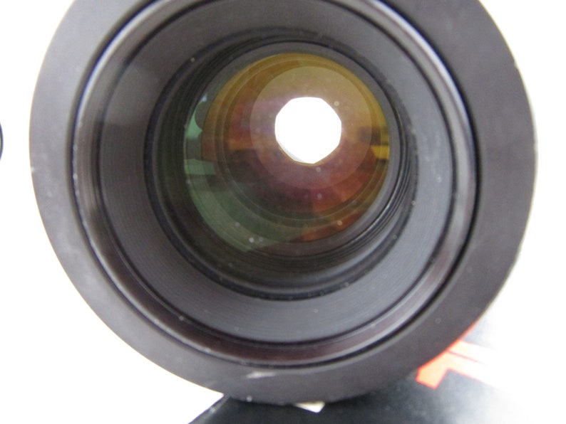 May include: Close-up of a black camera lens with a circular aperture. The lens has multiple layers of glass, visible through the aperture.