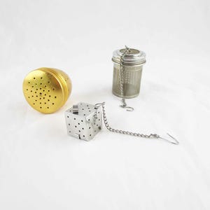 May include: Three tea infusers on a white surface. A gold-colored spherical infuser with small holes, a silver cylindrical infuser, and a silver cube-shaped infuser with a chain and hook.