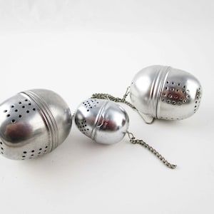 May include: Three silver-colored metal tea infusers of varying sizes, each featuring a spherical shape with small circular perforations. The infusers are connected by a thin metal chain, and each piece has a central seam for opening and closing.