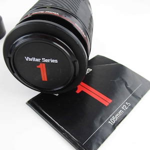 May include: A black Vivitar Series 1 105mm f/2.5 lens with a red and white label. The lens is sitting on a black instruction booklet.