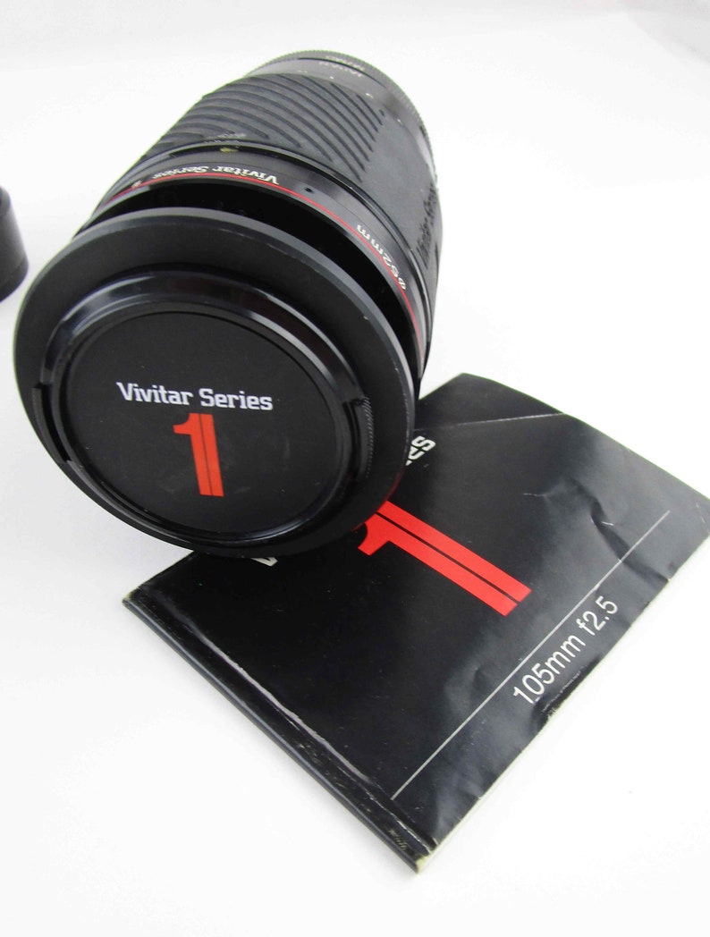 May include: A black Vivitar Series 1 105mm f/2.5 lens with a red and white label. The lens is sitting on a black instruction booklet.