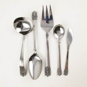 May include: A set of five silver-plated serving utensils, including a gravy ladle, a serving spoon, a serving fork, a butter knife, and a small spoon. The utensils have ornate handles with a floral design.