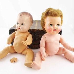 May include: Two vintage baby dolls with painted features and open-mouthed expressions. One doll has brown hair, the other has blonde hair. One doll is missing a hand. The dolls are sitting in front of a vintage suitcase.