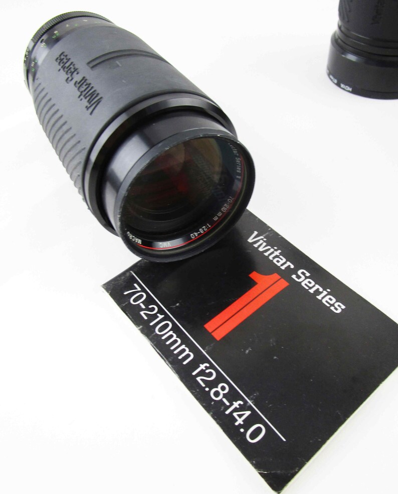 May include: A black Vivitar Series 70-210mm f2.8-f4.0 zoom lens with a black lens cap. The lens is sitting on a black and white instruction manual.