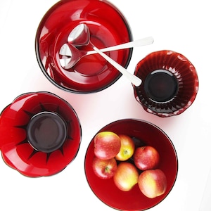 May include: An overhead shot showcases a collection of red lacquerware bowls and serving spoons. The bowls vary in size and shape, some with decorative edges. One bowl contains apples. The spoons are silver.