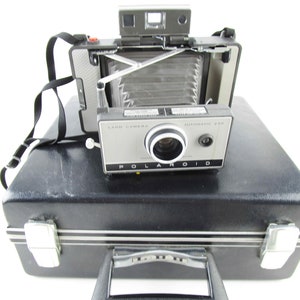 May include: A vintage Polaroid Land Camera Automatic 230 in a black carrying case. The camera is silver and black with a lens and a viewfinder. The case has a handle and two latches.