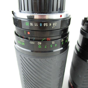 May include: A black camera lens with a silver ring and a depth of field scale marked in feet and meters. The lens has a black rubberized grip and the text "Tokina" is visible on the side.