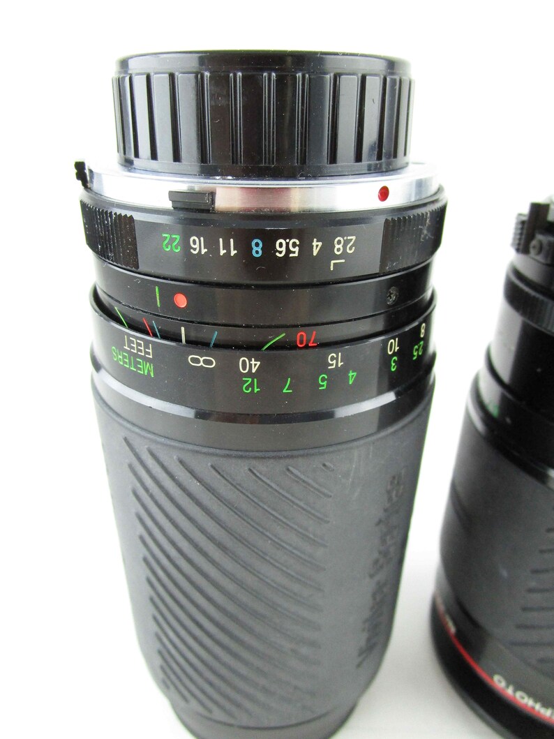 May include: A black camera lens with a silver ring and a depth of field scale marked in feet and meters. The lens has a black rubberized grip and the text "Tokina" is visible on the side.