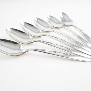 May include: Six silver spoons arranged on a white surface. The spoons have a classic design with a rounded bowl and a slender handle. The handles are decorated with a delicate floral pattern. The spoons are reflective, showing the light.
