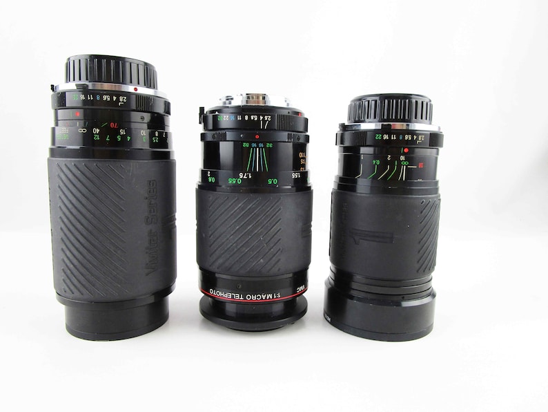 May include: Three black camera lenses with silver accents. The lenses have markings in green and red. The lens on the left has the text "Vuiker Series" printed on the side. The lens in the middle has the text "SIMACRO TELEPHOTO" printed on the side.