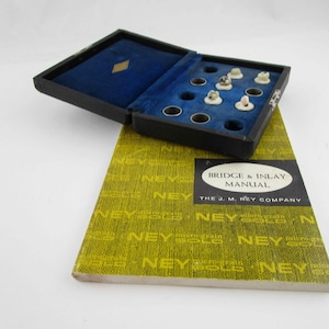 May include: An open black box with a blue velvet interior holds several small, off-white objects. The box sits atop a yellow and green book with the text "BRIDGE & INLAY MANUAL" and "THE J. M. NEY COMPANY".