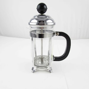May include: A clear glass French press coffee maker with a stainless steel frame and lid. It has a black handle and a black knob on top. The base has the word "Bonjour" printed on it.