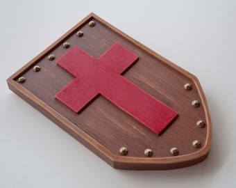 Puede incluir: Un escudo de madera con una cruz roja en el centro. El escudo está decorado con tachuelas de metal dorado alrededor del borde.