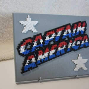 May include: A gray canvas with a red, white, and blue beaded design that says "CAPTAIN AMERICA". There are two white beaded stars on the canvas.