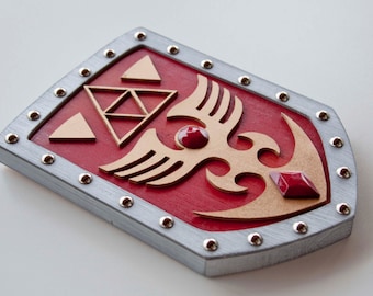 May include: A red wooden shield with a gold design featuring a stylised bird with wings and a triangular pattern. The shield is decorated with silver studs around the perimeter and has two red gemstones.